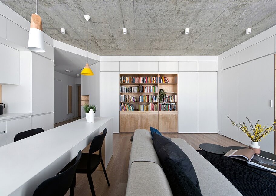 Phòng khách hiện đại với sofa và kệ sách, nội thất tối giản, trần bê tông, đèn treo độc đáo.