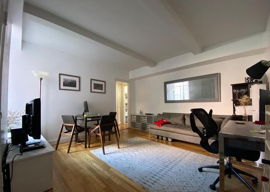 Phòng khách hiện đại, sáng sủa với bàn làm việc, sofa xám và sàn gỗ sáng. Có tranh trang trí và đèn đứng.