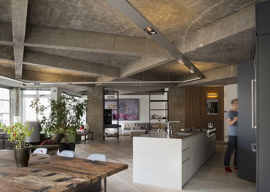 Phòng khách hiện đại với trần bê tông, bếp mở và cây xanh. Không gian rộng thoáng và nội thất sang trọng.