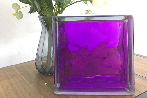 Khối thủy tinh màu tím sáng bóng đặt trên bàn gỗ cạnh bình hoa lá xanh.