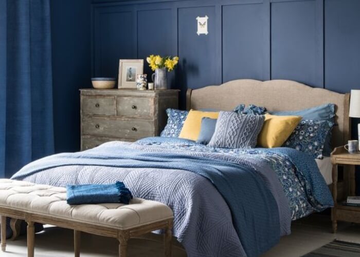Phòng ngủ thanh lịch với tường xanh navy, giường ấm cúng, chăn xanh lam, gối vàng, tủ gỗ và bình hoa trên đầu giường.